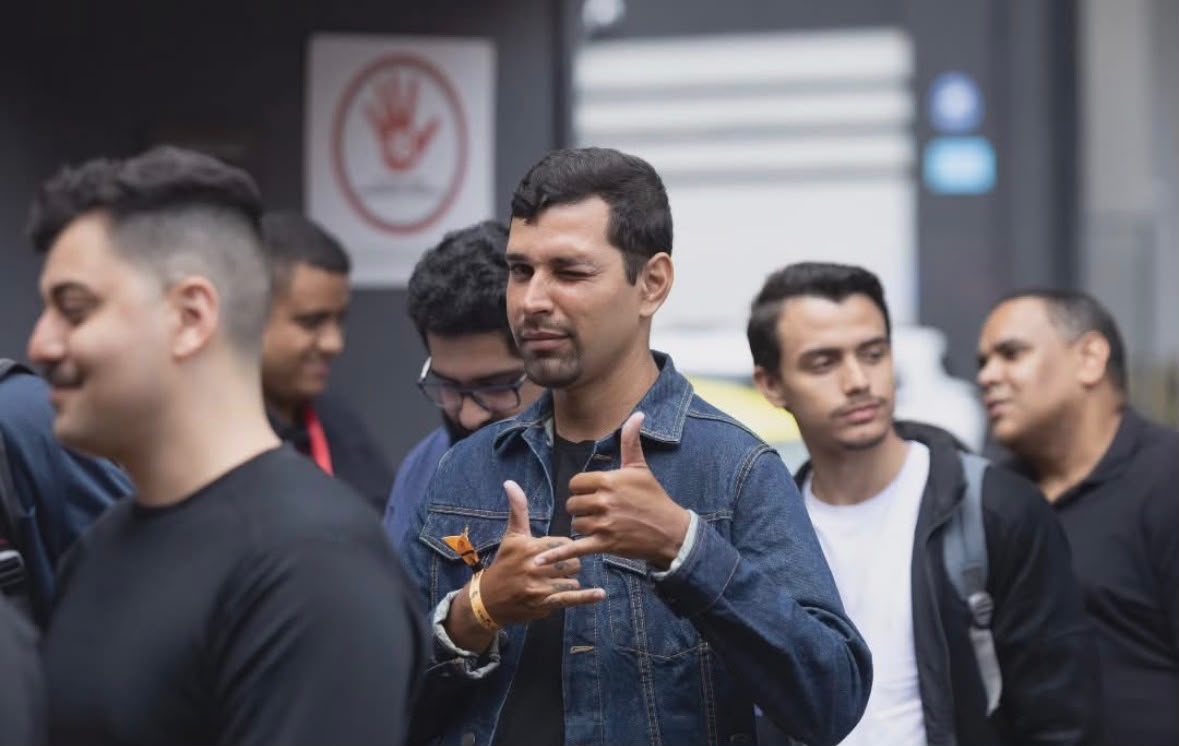 Derek Canarin em evento público, de jaqueta jeans, entre outras pessoas.